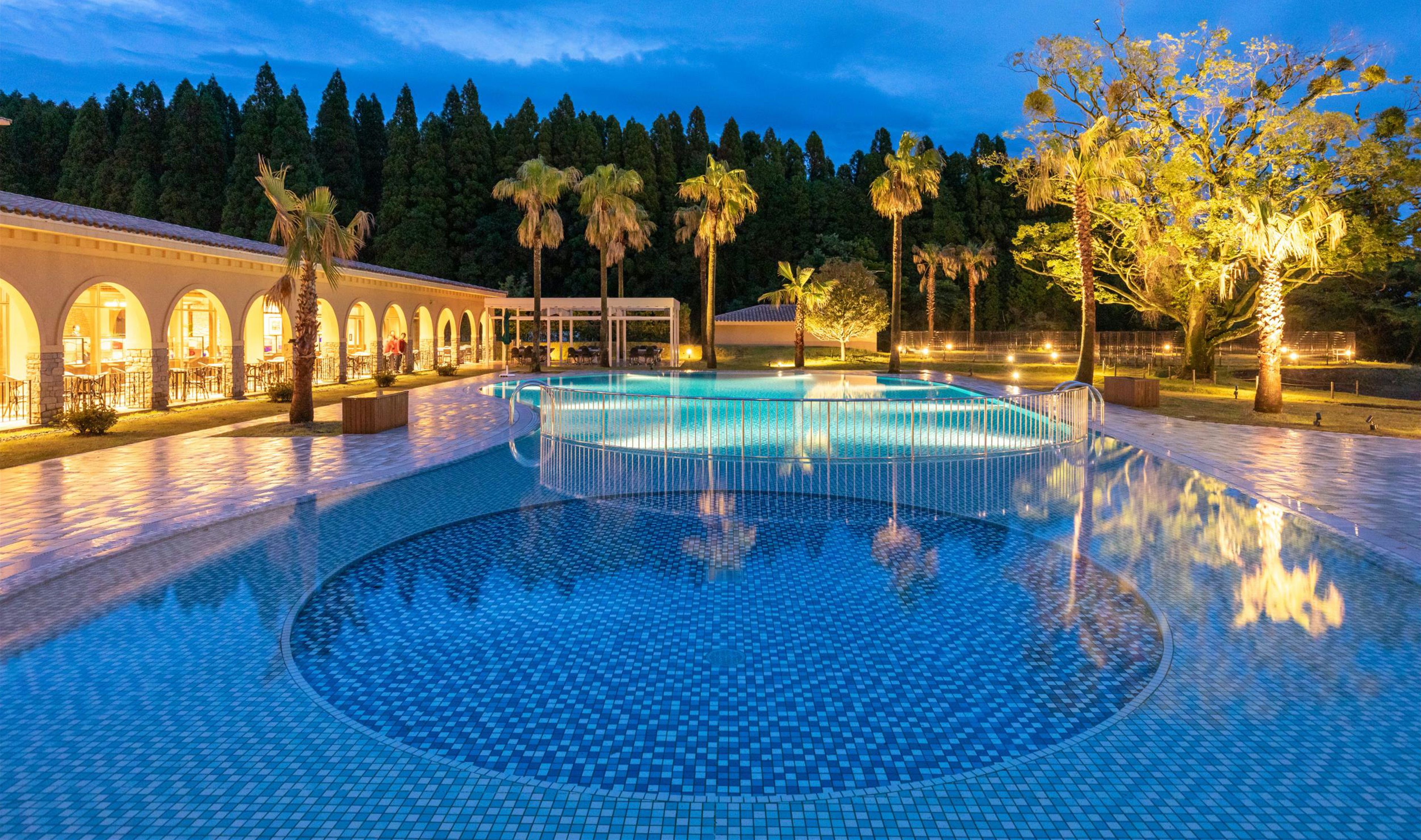 Zona de piscina con suelo de baldosas y un edificio con ventanas de arco al fondo.