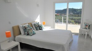 A bedroom with a large bed, white nightstands, and a view of a balcony with a fence and greenery.