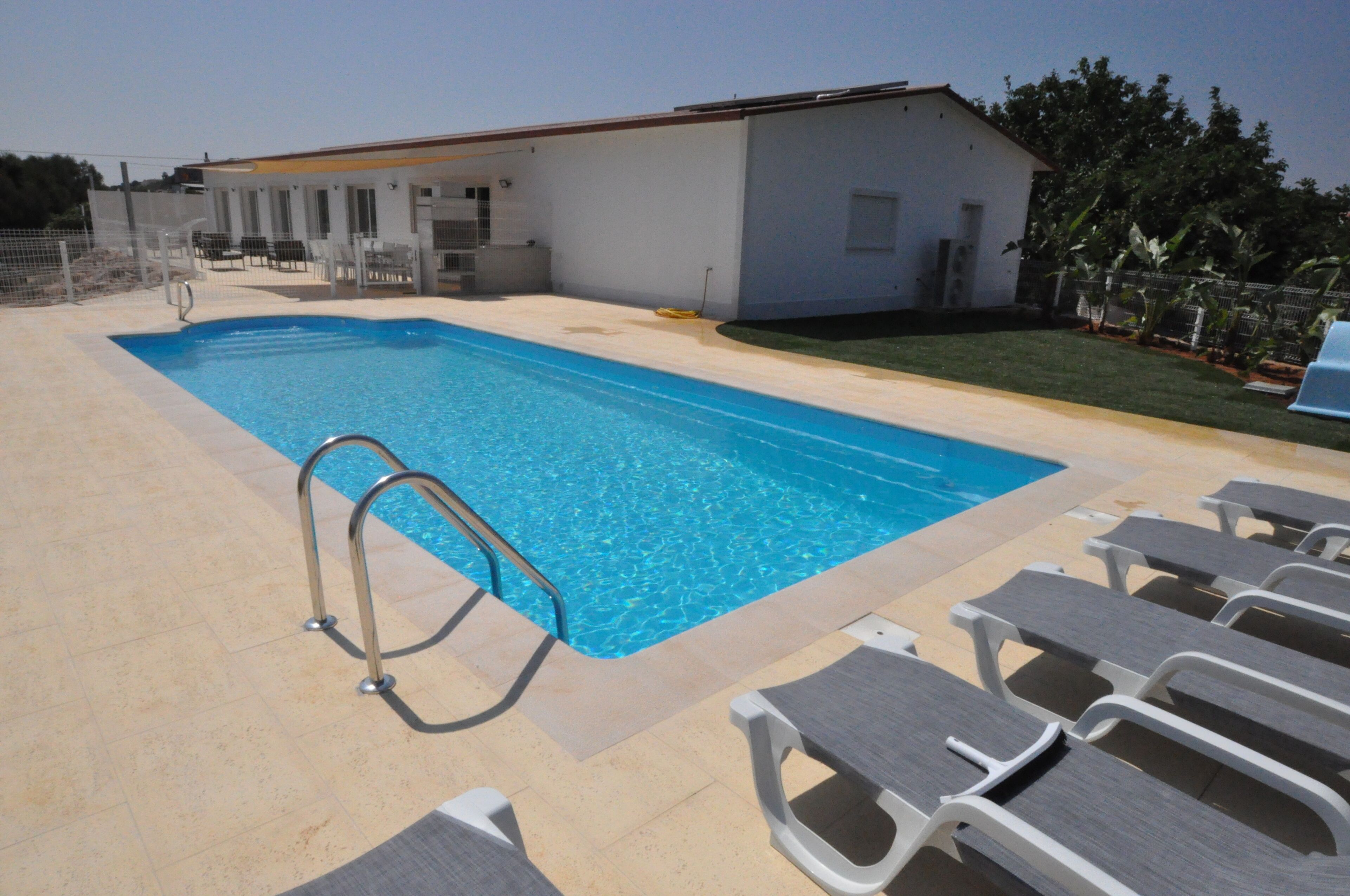 Une piscine rectangulaire avec un escalier en métal, entourée de chaises longues et bordée d’un bâtiment à l’extérieur blanc.