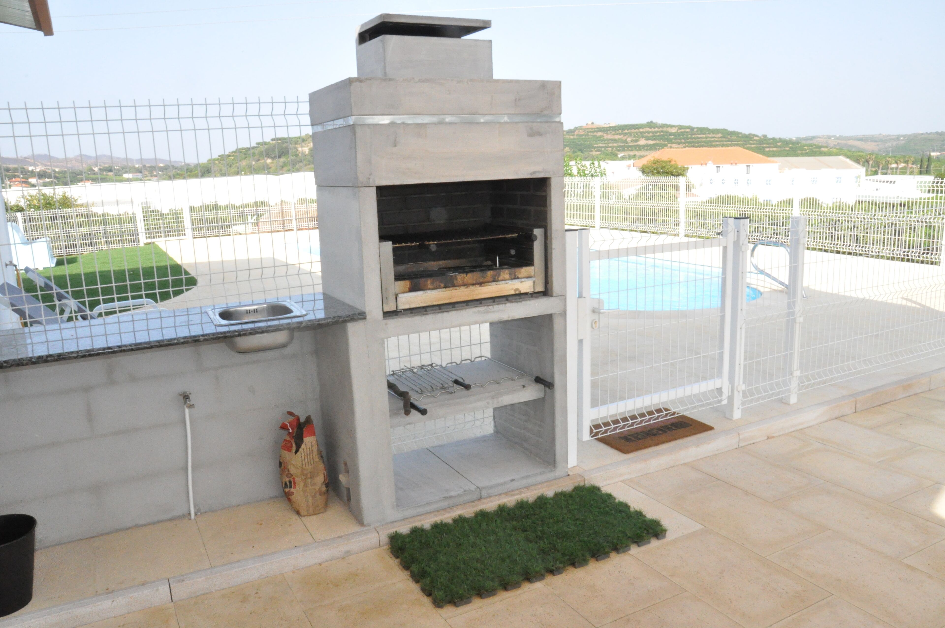 A concrete outdoor kitchen with a sink and a grill, surrounded by a white fence and a pool.
