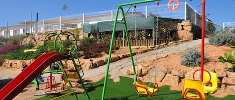 A playground with swings and slides, surrounded by rocks and plants.