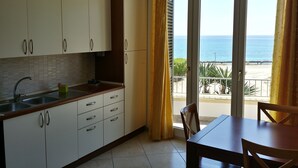 Una cocina con armarios blancos, un lavamanos y una zona de comedor con mesa y sillas. Se ve el mar a través de las puertas correderas de vidrio.