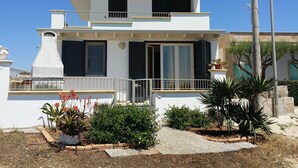 Un edificio de dos pisos con balcón, chimenea y un pequeño jardín con plantas y un sendero.