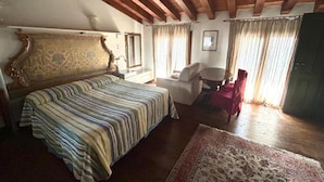 A bedroom with a large bed, a sofa, a small table, and a chair. There is a window with curtains, a mirror, and a door. - Damabianca junior (Porto Gruaro)