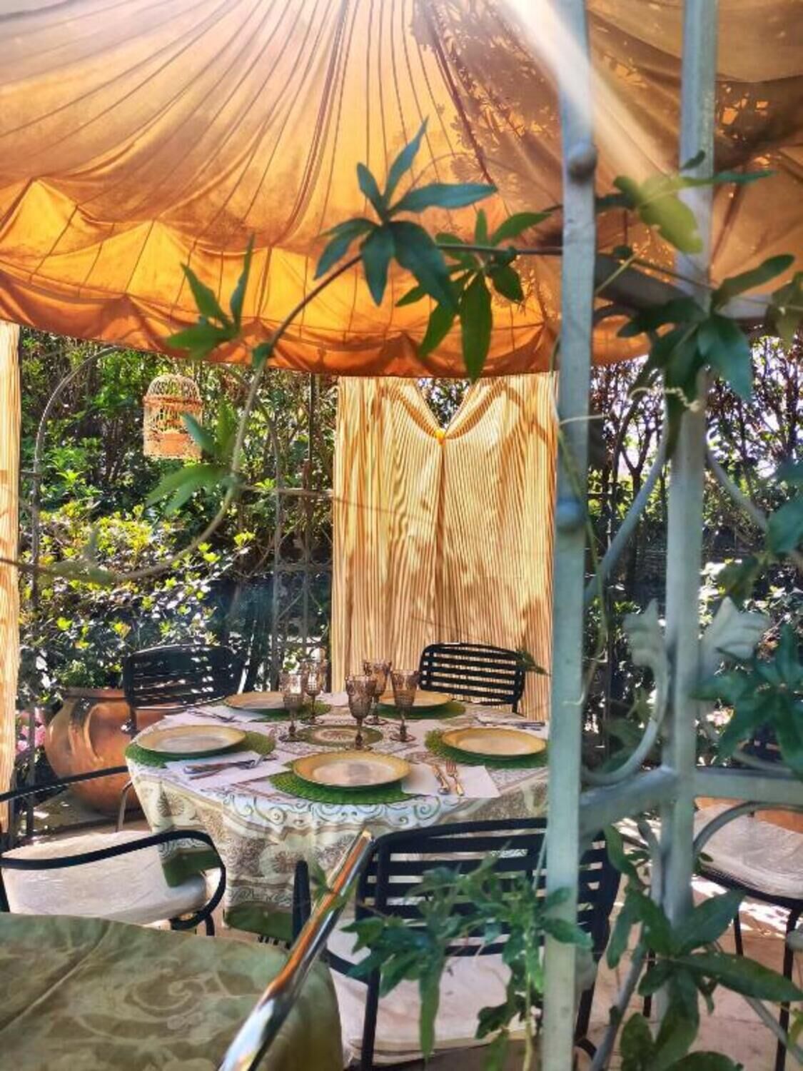 An outdoor dining area with a table set for two, surrounded by chairs, under a canopy.
