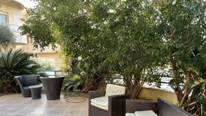 Un patio esterno con mobili in vimini, tra cui un divano e poltrone, immerso nel verde con vista su un edificio sullo sfondo.