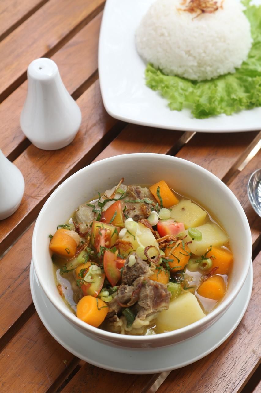 A bowl of stew with vegetables and meat, a plate of white rice, and two salt shakers on a wooden table.