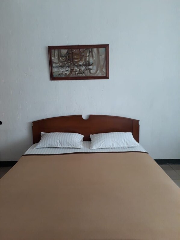 Una cama individual con cabecera de madera, dos almohadas y una colcha marrón en una habitación con pared blanca lisa y un cuadro abstracto enmarcado sobre la cama.