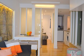 A modern living room with a sofa, a dining table with fruit, a kitchen with a washing machine, and a laundry basket.