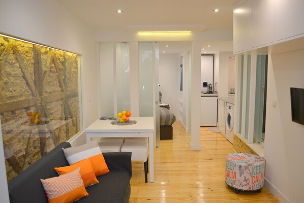 A modern living room with a dark sofa, a small table with fruits, and a colorful ottoman.