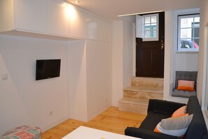 Uma sala de estar moderna com uma TV de tela plana na parede, um sofá com almofadas, um banquinho redondo pequeno e uma escada que leva a um andar superior.