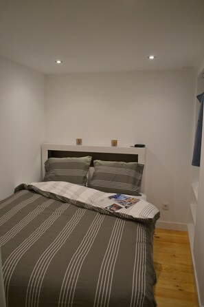 Quarto pequeno e bem organizado com uma cama de solteiro, cabeceira e uma mesinha de cabeceira com abajur.