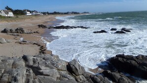 Une plage rocheuse où les vagues se brisent sur le rivage, un ciel dégagé et des maisons en arrière-plan.
