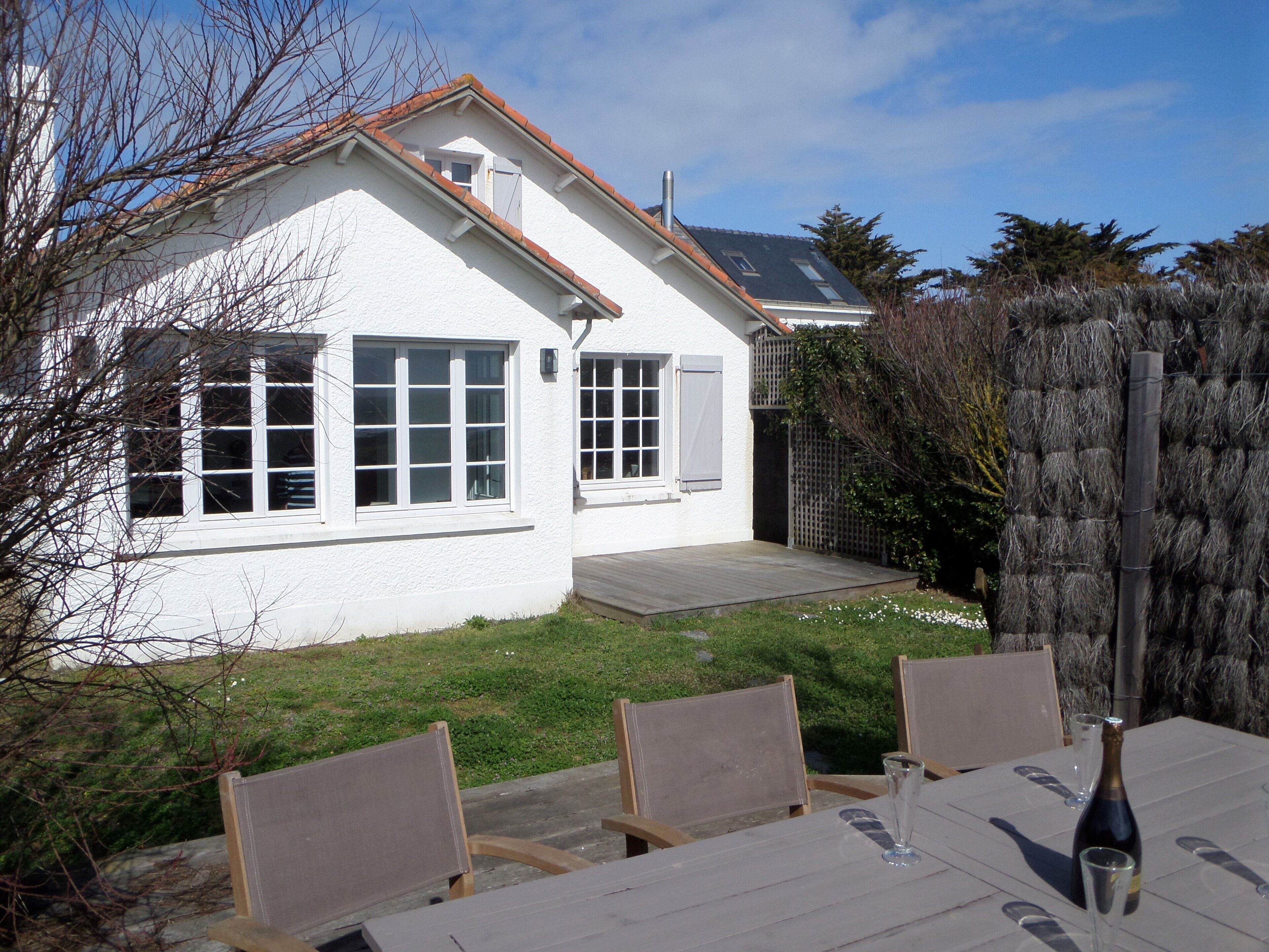 Une maison blanche avec un toit en pente, une cheminée bien visible et une petite fenêtre avec un volet. Devant la maison, on trouve une terrasse en bois, ainsi qu’une table avec des chaises et une bouteille de champagne.