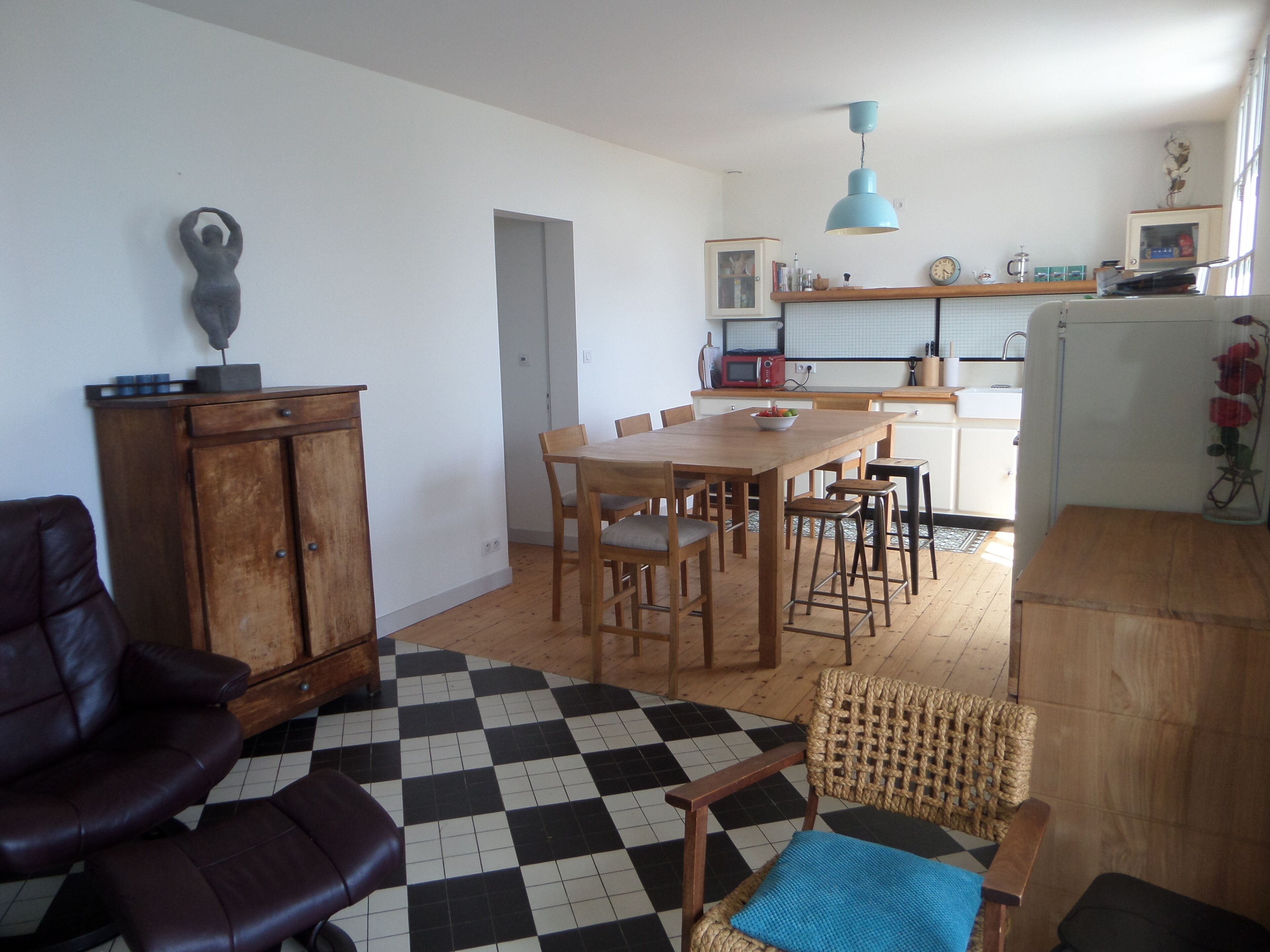 Uma sala de jantar com piso xadrez, móveis de madeira e uma cozinha ao fundo.