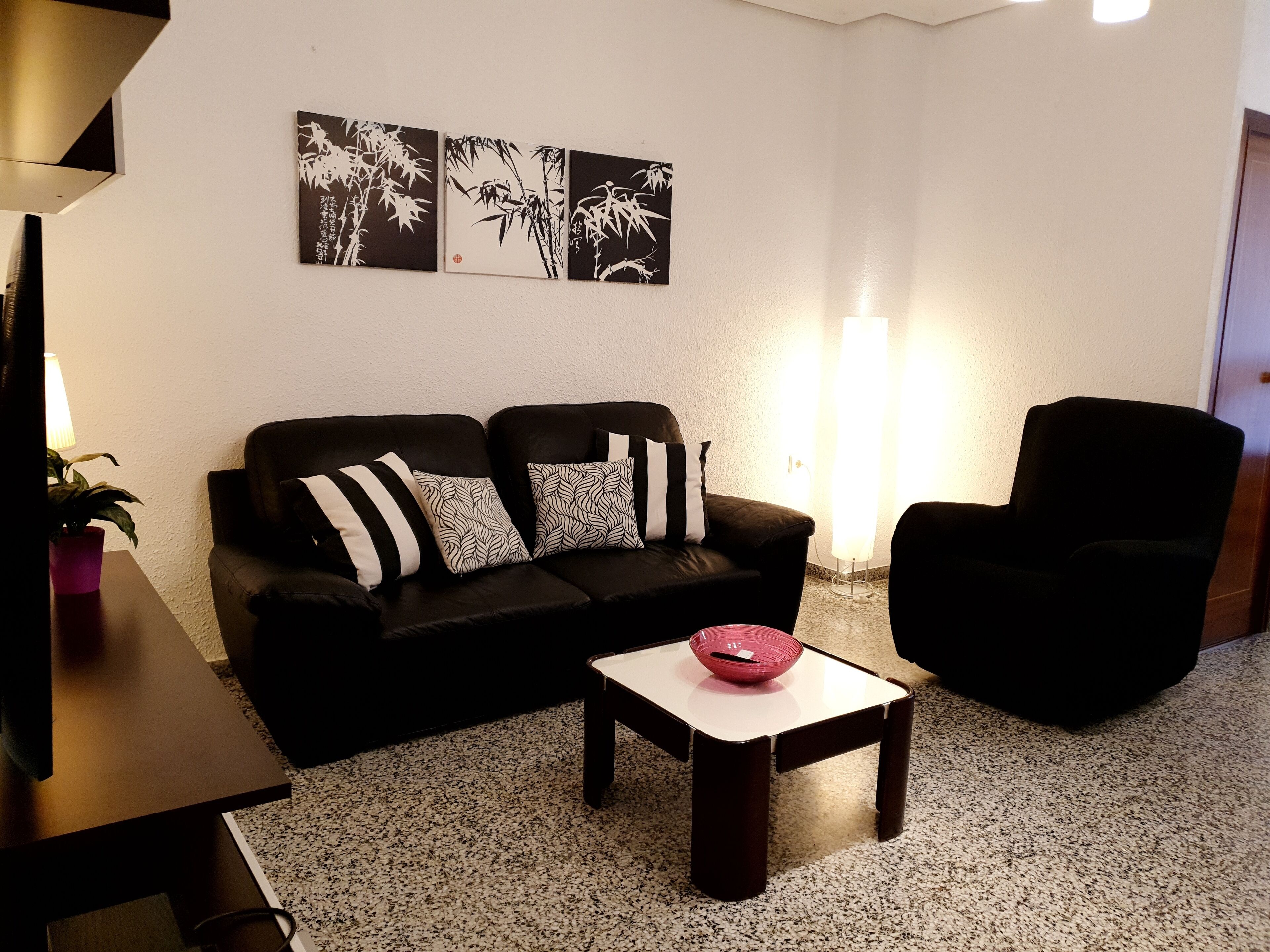 A living room with a black sofa, a small table with a red bowl, and a black armchair. There are three wall-mounted paintings above the sofa.