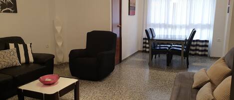 Un salon avec un canapé noir, un fauteuil noir, une table à manger avec des chaises et un bol rouge posé sur une table basse.