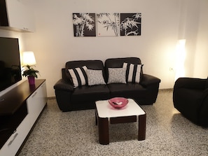 Un salon avec un canapé noir, deux coussins à motifs, une petite table blanche sur laquelle se trouve un bol rose, et un téléviseur posé sur un meuble.