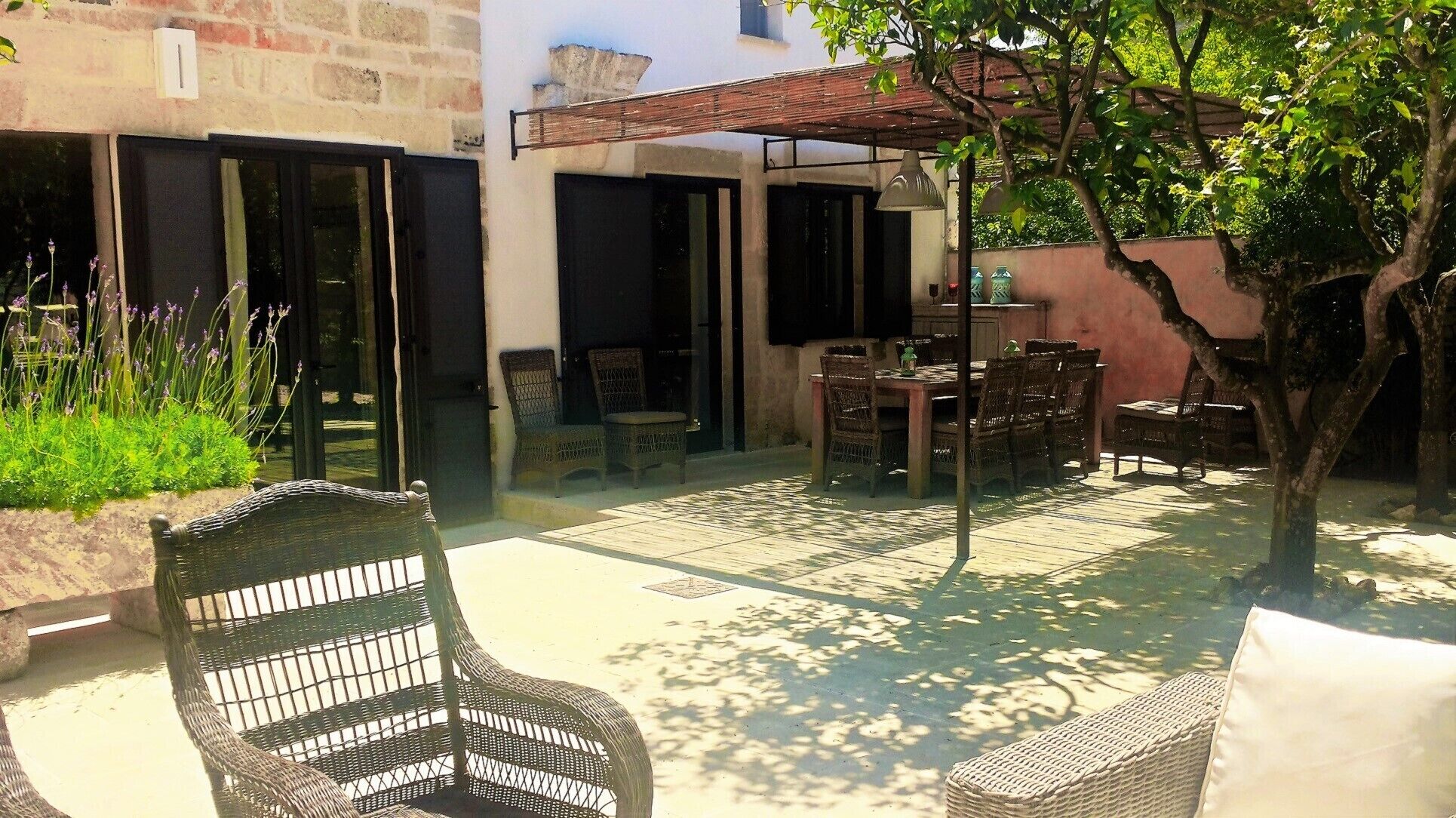 Un patio con mobiliario de mimbre, una mesa y sillas. Hay una pérgola que da sombra y se ve un árbol.