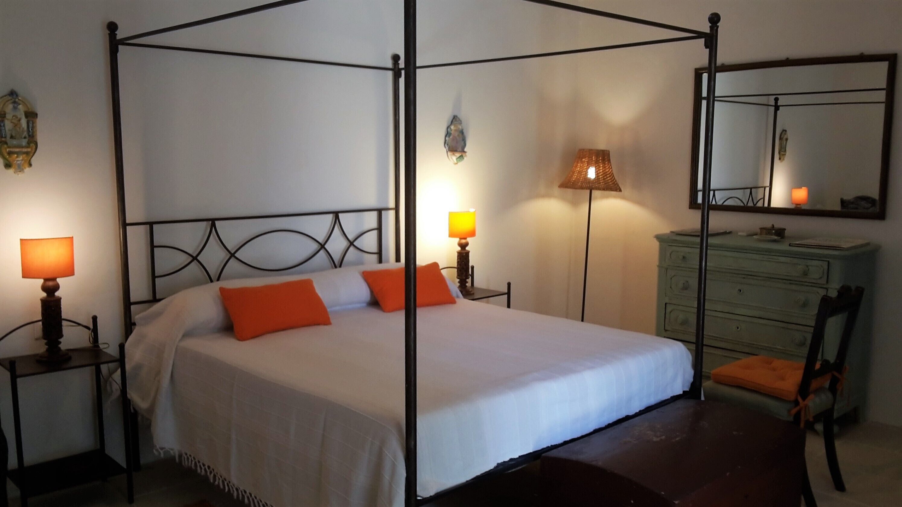 Uma cama com dossel com duas almofadas, uma mesa de cabeceira com abajur, uma cômoda e um espelho na parede.