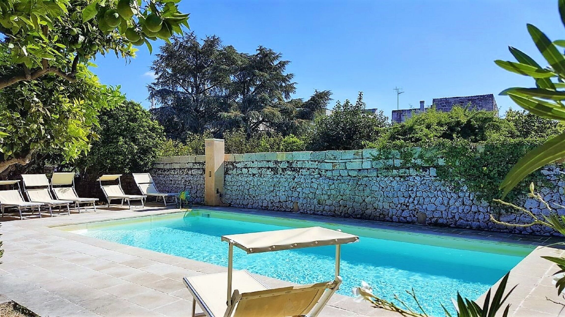 Ein Schwimmbecken mit Liegestühlen, einer Steinmauer und Bäumen im Hintergrund.