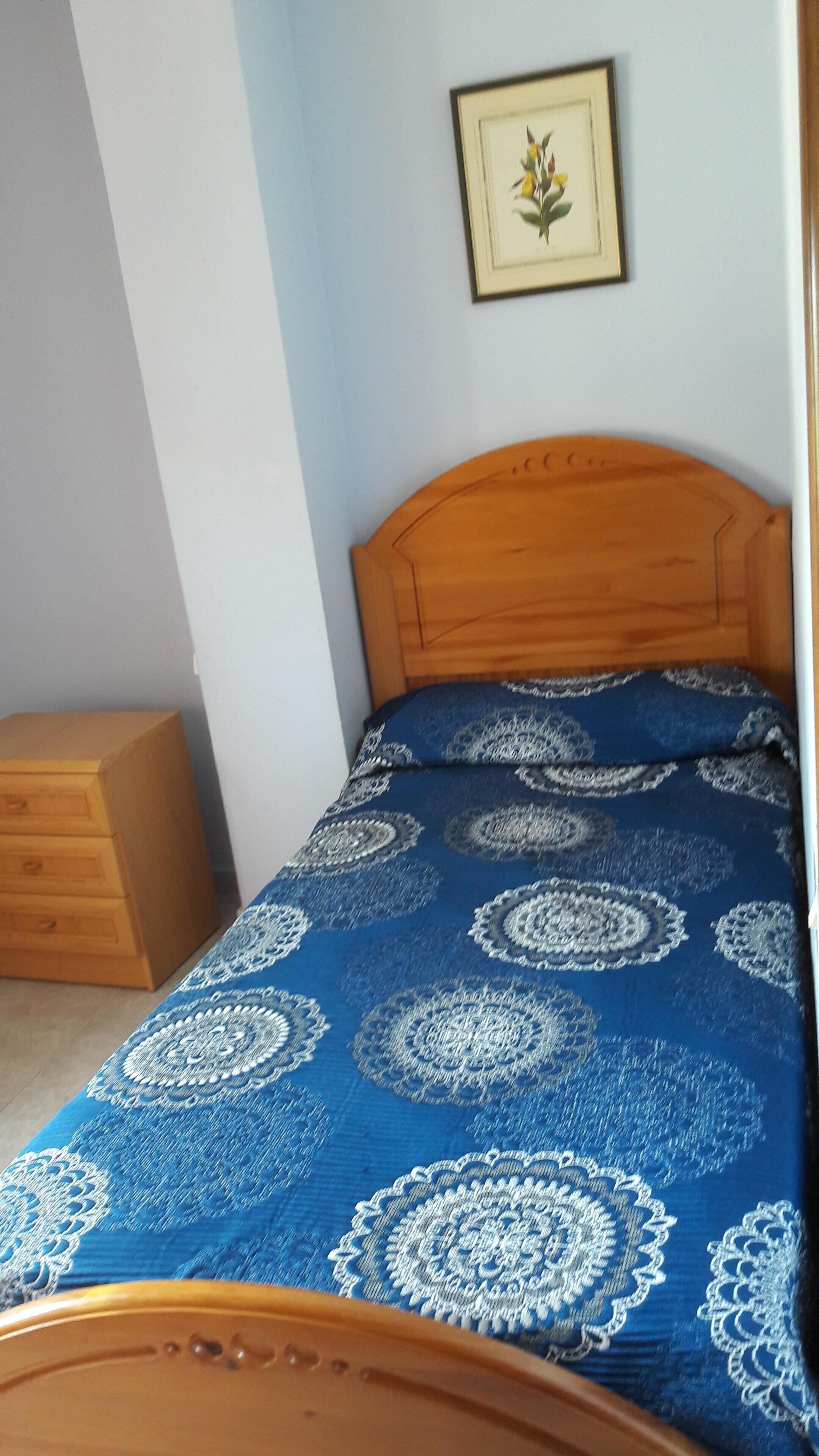 Ein Schlafzimmer mit einem Holz-Kopfteil, einer blau gemusterten Tagesdecke, einem Nachttisch aus Holz und einem gerahmten Bild an der Wand.