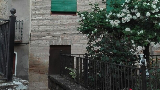 Ein mehrstöckiges Gebäude mit grünen Fensterläden, einem Balkon im zweiten Stock und einer kleinen Gartenanlage mit weißen Blumen.