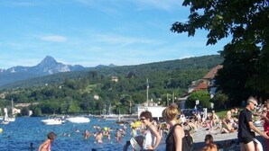 A crowded beach with people swimming and sunbathing, boats in the water, and a mountain in the background.