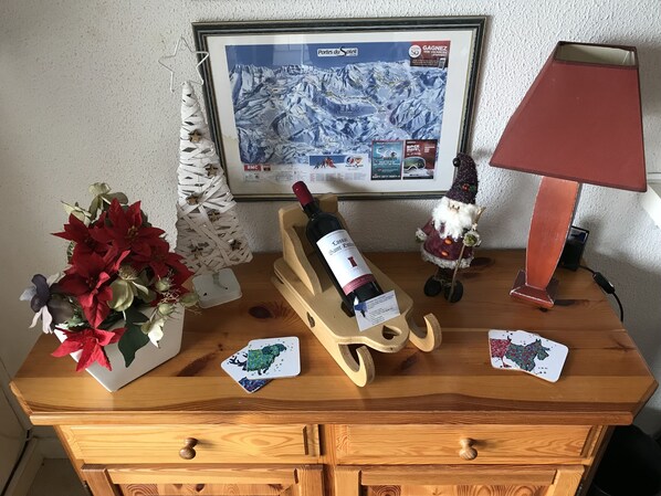 Una mesa de madera con un mapa enmarcado, una botella de vino, un pequeño árbol de Navidad, una lámpara y un juego de cartas.
