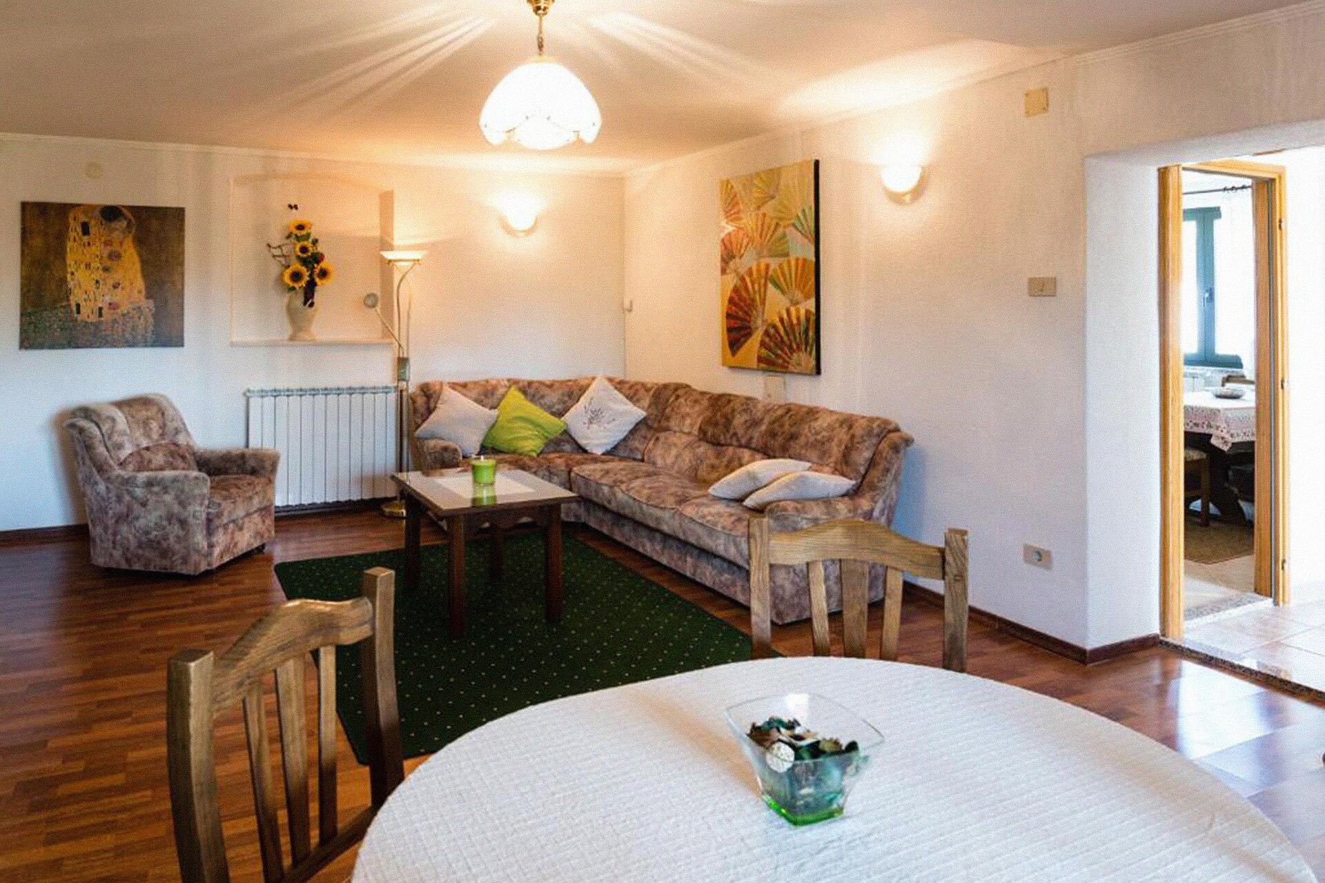 A cozy living room with a sofa, armchairs, a coffee table, and a dining table. There are paintings on the walls and a radiator below the sofa.
