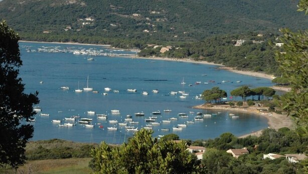 Vista costera con barcos en el agua, una playa y un promontorio cubierto de bosque.