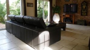 Una sala de estar con un sofá de cuero negro, un televisor en una pared de ladrillo y un gran ventanal con cortinas.