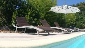 Piscina con sillas de descanso y sombrillas, rodeada de árboles y un edificio con techo de tejas.