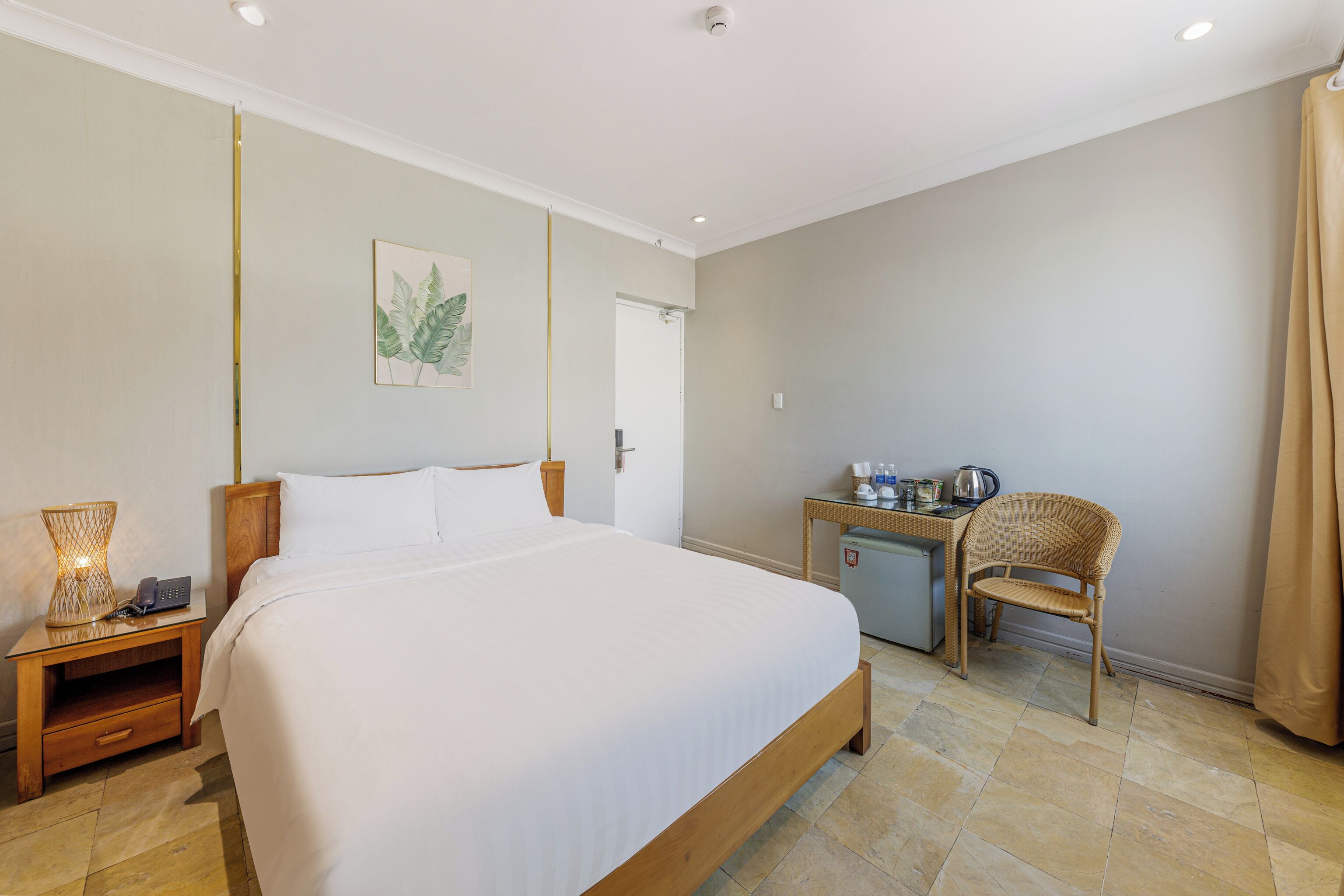 A hotel room with a large bed, bedside table with a lamp, a small desk with a chair, and a door leading to another room.
