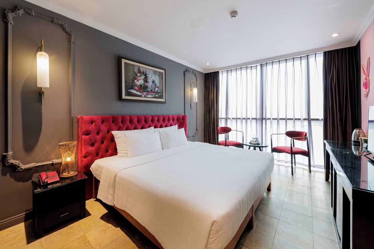 A hotel room with a large bed, a red headboard, a painting on the wall, a desk with a chair, and a large window with curtains.