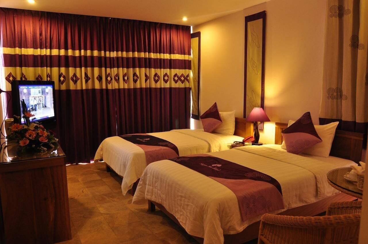 A hotel room with two beds, a desk with a computer and flowers, and a small table with a lamp.