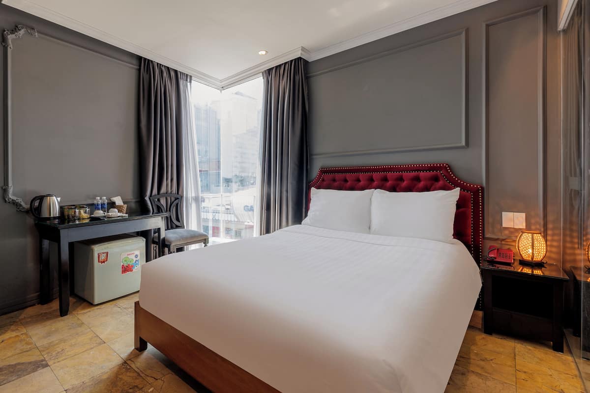 A hotel room with a large bed, a desk with a chair, a small refrigerator, and a nightstand with a lamp.