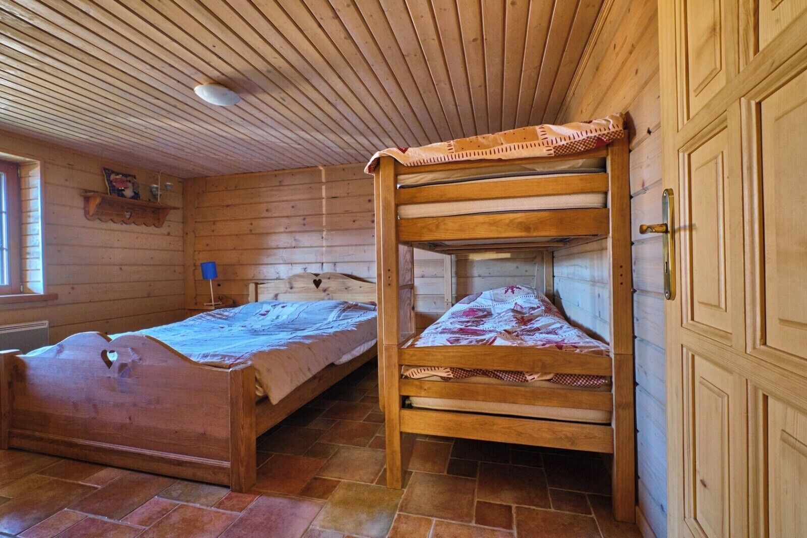 Une chambre dans un chalet en bois, avec deux lits superposés, une fenêtre avec des stores et une petite étagère ornée d’objets décoratifs.