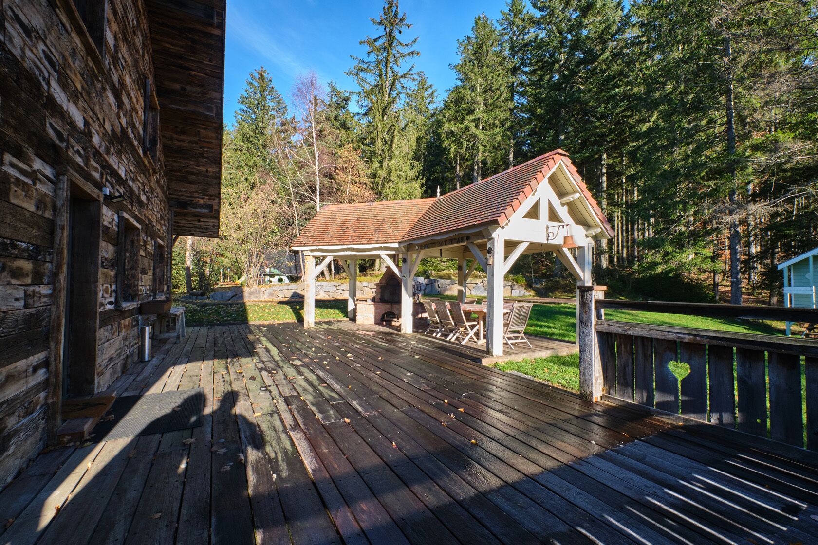 Eine Holzterrasse mit überdachter Sitzgruppe und einem Kamin aus Stein.