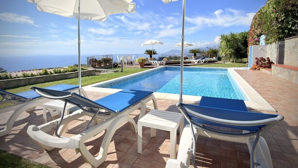 Zona de piscina con sillones de playa y sombrillas, vista despejada al océano y jardín cuidado.