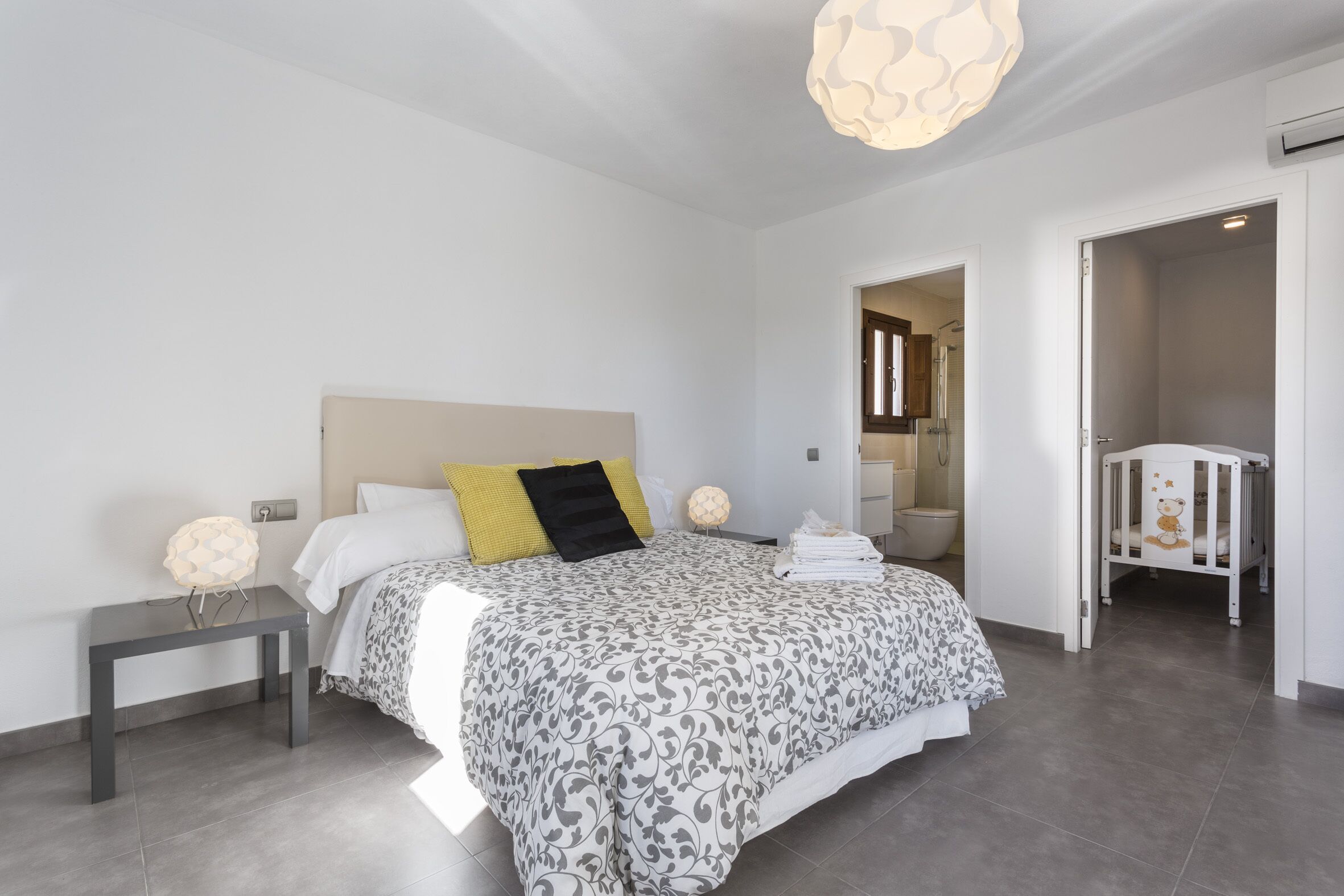 Quarto moderno com uma cama grande, mesas de cabeceira, um criado-mudo, berço e um banheiro visível por uma porta aberta.