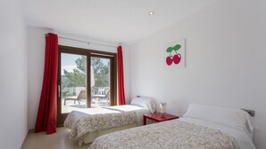 A hotel room with two beds, a window with red curtains, a small table, and a wall-mounted picture of cherries.
