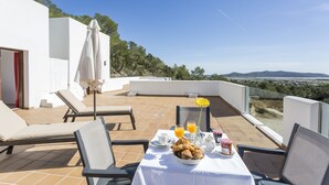 A terrace with a table set for breakfast, including croissants, juice, and coffee, overlooking a scenic landscape.
