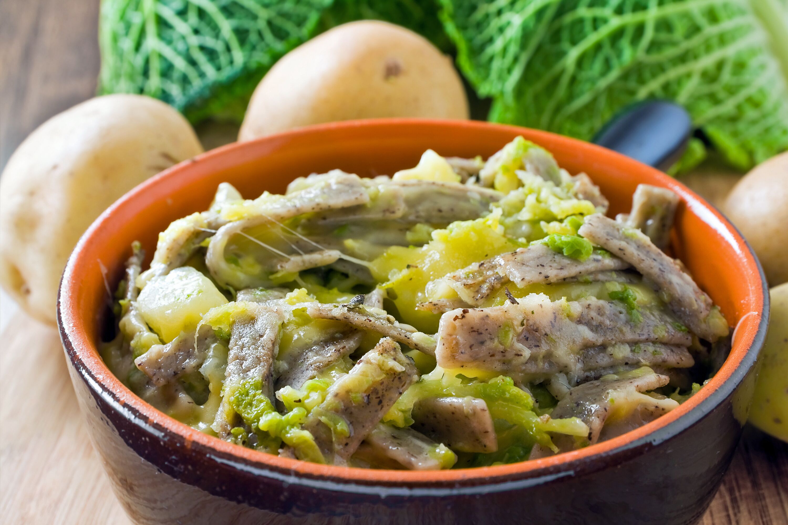 Eine Schüssel mit Eintopf, Fleisch und Gemüse, umgeben von Kartoffeln und Kohl.