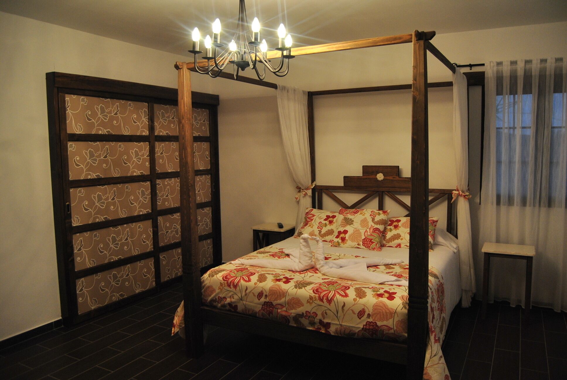 A four-poster bed with floral bedding, a chandelier, a wooden headboard, and a window with curtains.