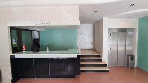 A reception area with a counter, a small desk, a telephone, and a sign above the counter. There is a staircase leading to an elevator. - Hotel Yasmine (Sfax)