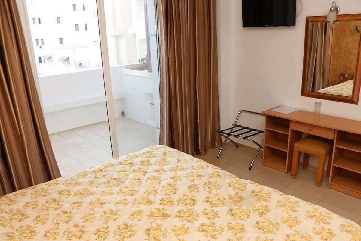 A hotel room with a bed, a desk, a chair, a mirror, and a view of the outside balcony.