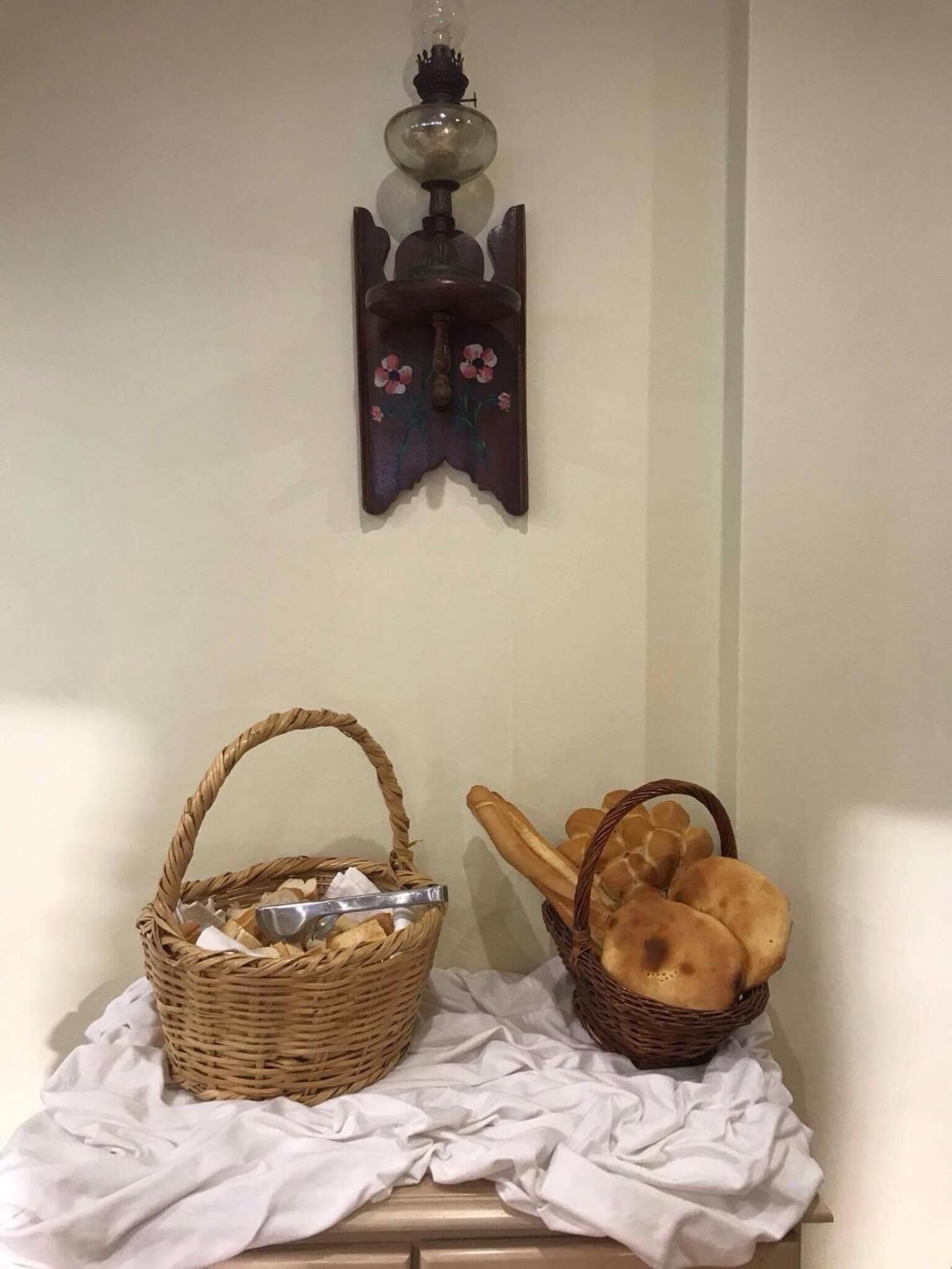 A wall-mounted lamp with floral design, a wicker basket with utensils, and another basket with bread.