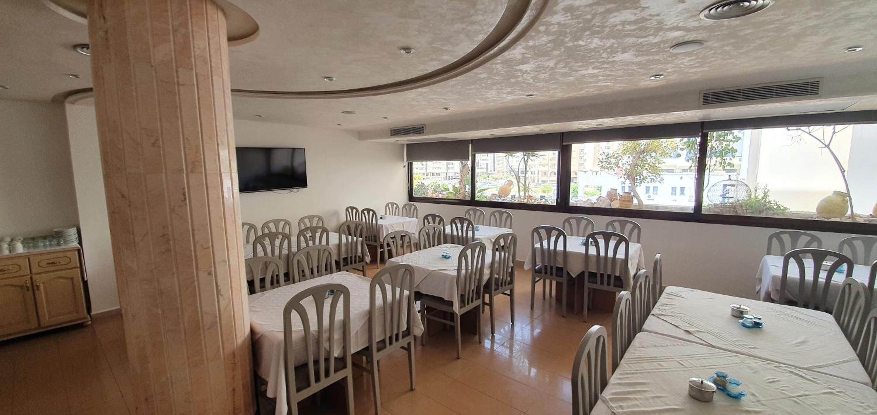 Un comedor con varias mesas y sillas, un televisor montado en la pared y ventanas amplias con vistas al exterior.
