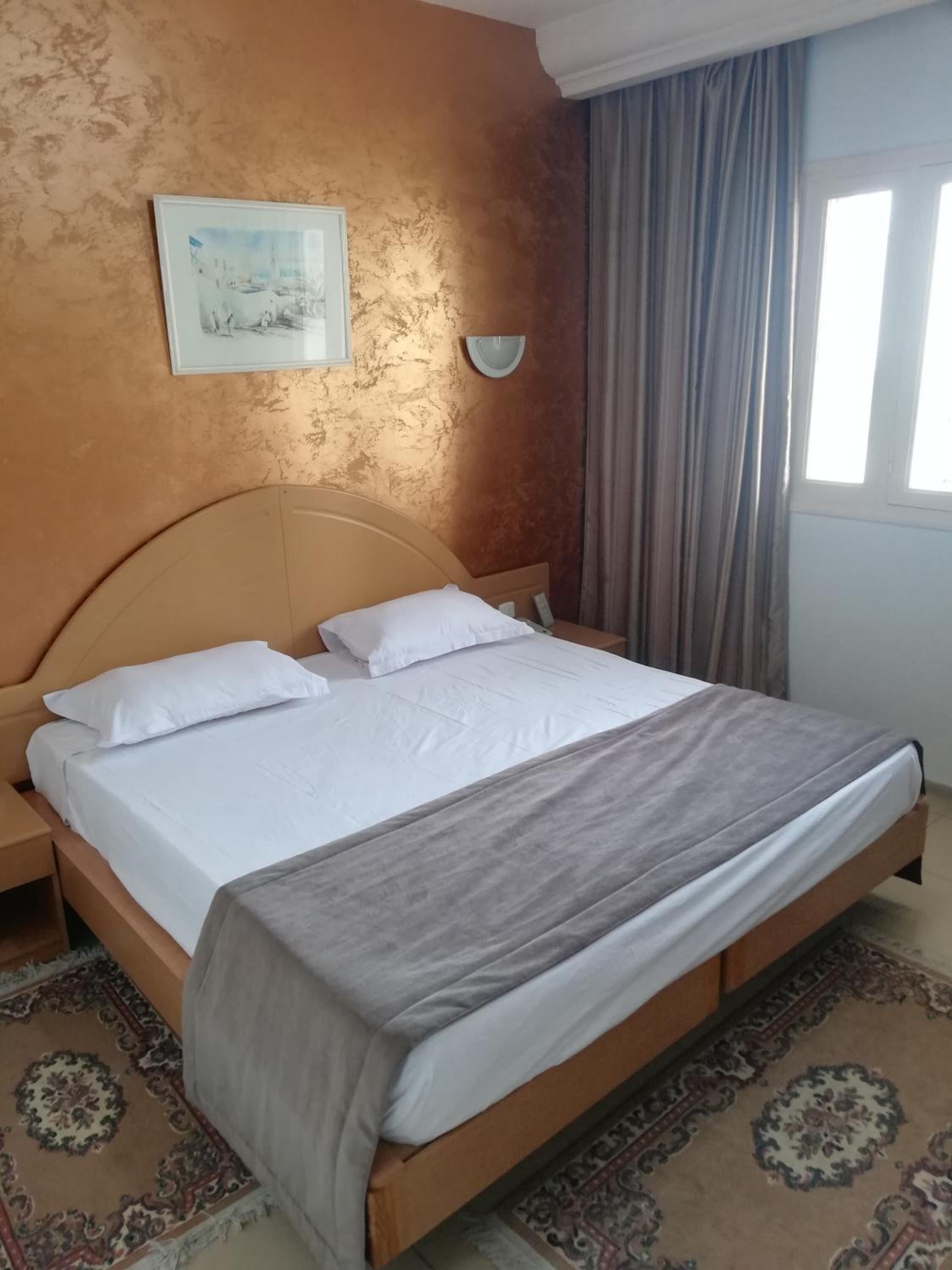 Un cuarto de hotel con una cama grande, una mesita de noche, una lámpara de pared, una ventana con cortinas y un cuadro en la pared.